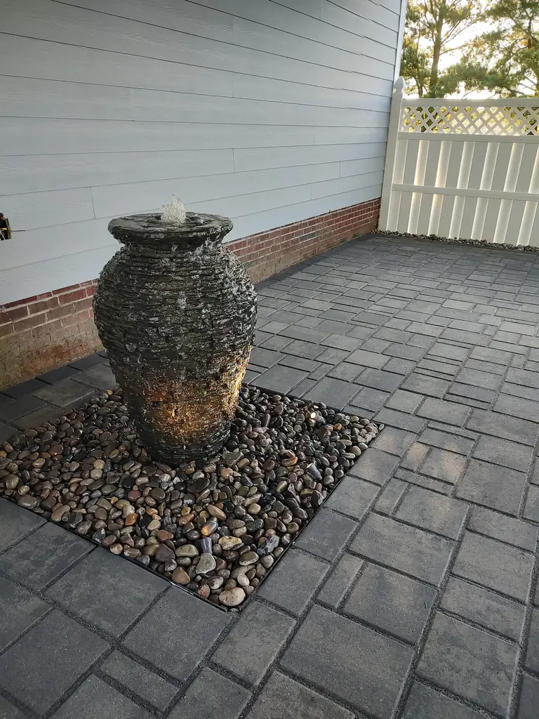 Outdoor living area with paver flooring and a stone urn fountain beside a white fence